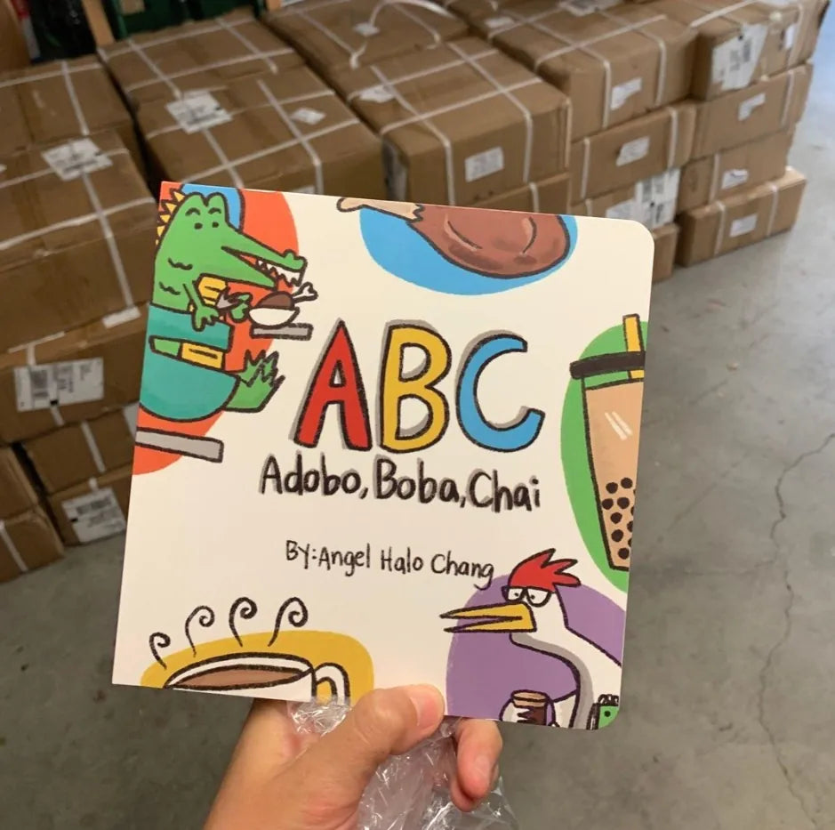 A copy of ABC Adobo Boba Chai by Angel Halo Chang held up in front of boxes from the printer